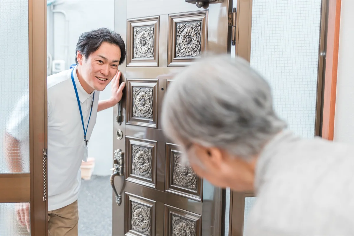 玄関で高齢の利用者に笑顔であいさつする男性スタッフ