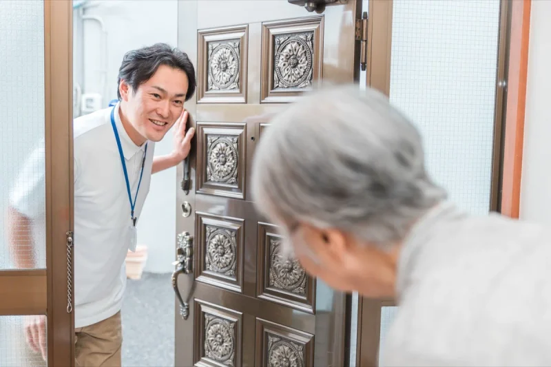 玄関の扉を開け、高齢の利用者に笑顔であいさつする男性スタッフ